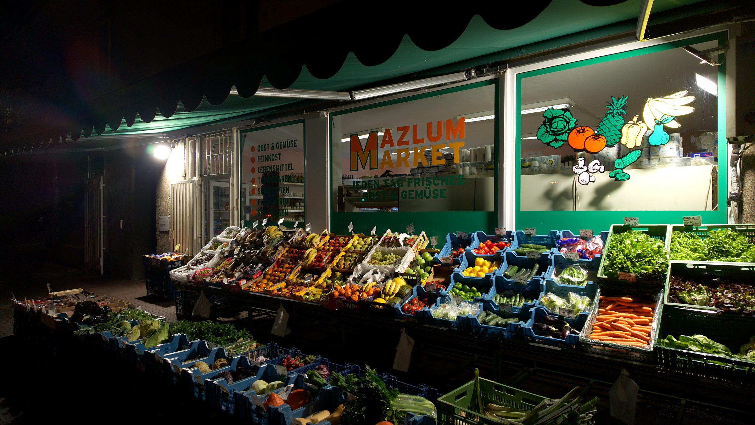 Mazlum Market am Abend: die grüne Markise, das Schild und die Obst- und Gemüseauslage vor dem Laden